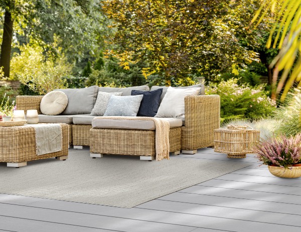 Outdoor patio with wicker sectional sofa and neutral cushions on a gray composite deck, surrounded by lush greenery and trees, creating a cozy seating area for warm weather relaxation.
