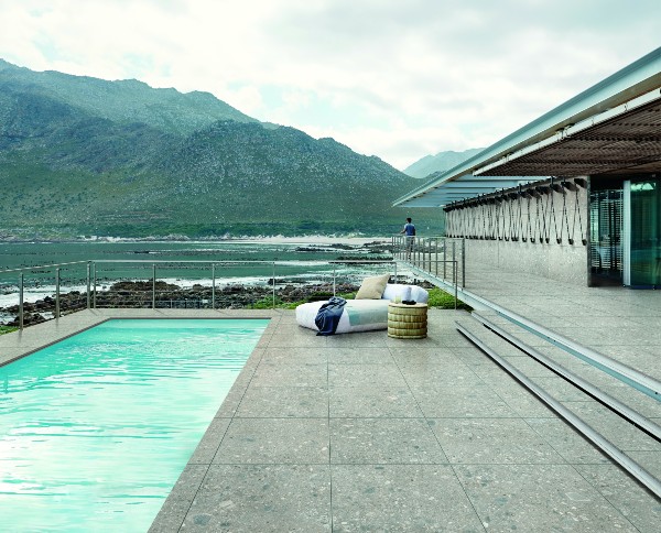 Modern poolside patio with light gray exterior tile overlooking the ocean and mountains, featuring minimalist outdoor seating and glass railing for an open, scenic view.