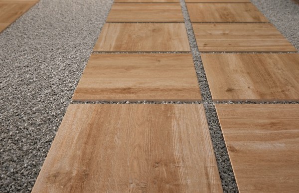 Outdoor walkway with square wood-look porcelain pavers set in gravel, leading to a modern patio area with outdoor dining and large potted plants.