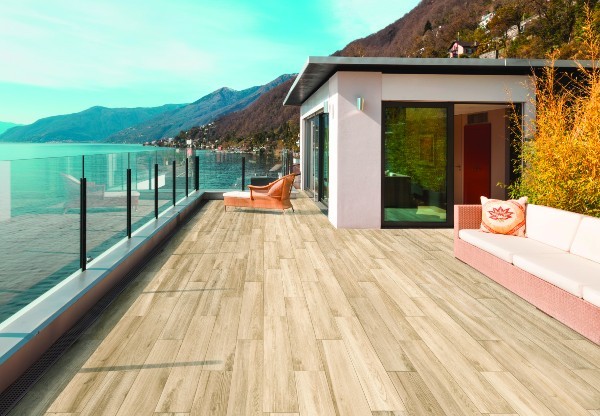 Rooftop patio with light wood-look porcelain tile flooring, modern outdoor seating, glass railing overlooking a lake, and mountains in the background under a bright blue sky.