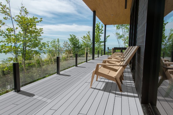 Modern outdoor deck with light gray composite decking, tan chairs, and glass railing overlooking trees and a lake under a clear sky.