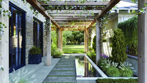 Outdoor walkway with large porcelain pavers surrounded by grass, a reflecting pool, and a wood pergola with climbing plants, leading to a green lawn.