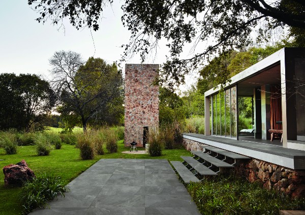 Modern home with large glass windows, outdoor porcelain paver patio, and a stone fireplace surrounded by lush greenery.