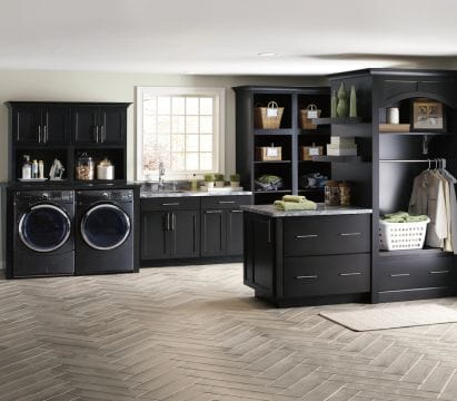 Stylish laundry room with dark cabinetry, herringbone flooring, shelving, and appliances under natural light.
