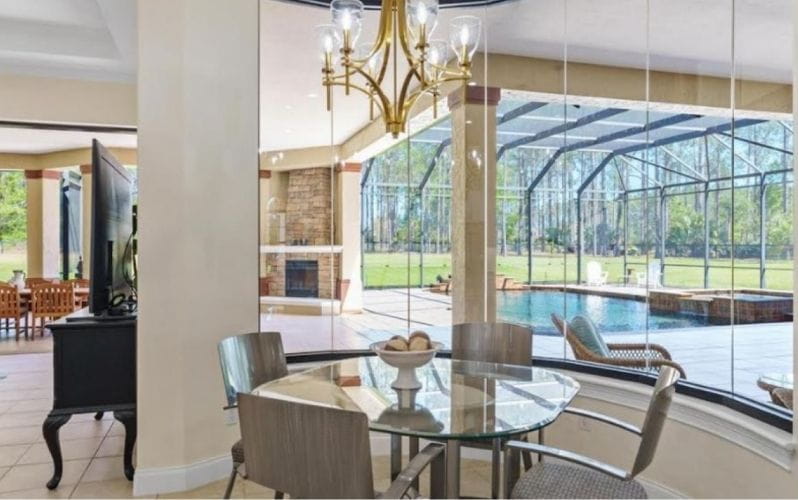 An interior photo featuring a dining area with a glass table, chairs, and a pool visible through large windows.