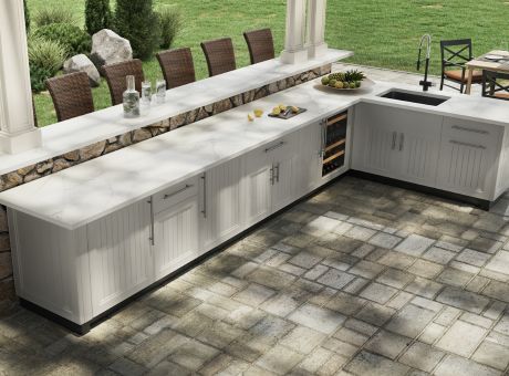 A stylish outdoor kitchen equipped with white cabinets, stone decorations, and seats nearby in a garden setting.