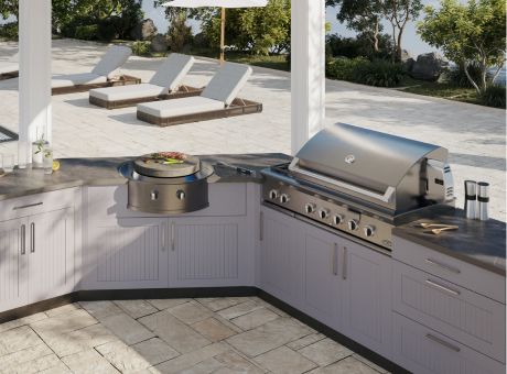 Outdoor kitchen setup with a barbecue grill and counter space on a patio with lounge chairs and greenery.