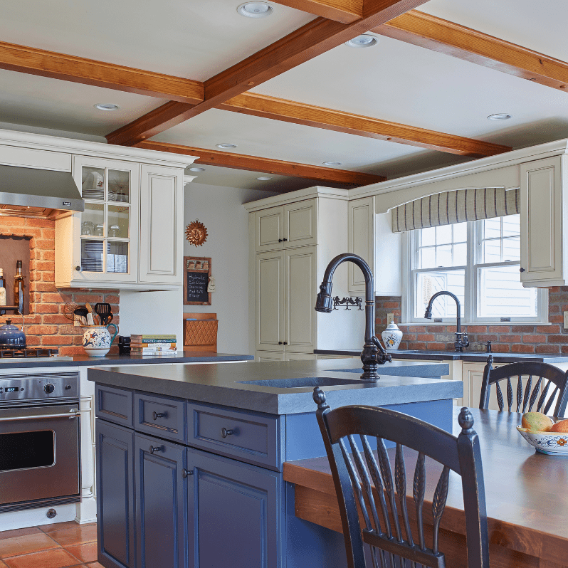 Exposed brick accents and wooden ceiling beams provide a rustic touch, while the vibrant blue kitchen island is topped with dark gray countertops, adding contrast and modern sophistication.