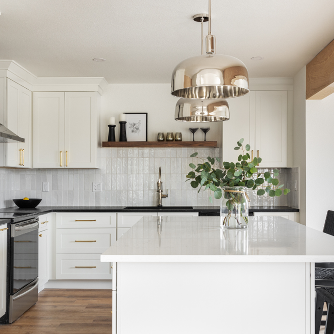 A modern kitchen with crisp white cabinetry, gold hardware, and sleek black appliances. The reflective pendant lights and warm wood accents create a balanced blend of contemporary and classic elements, while the soft tile backsplash adds subtle texture.