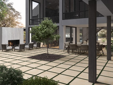 Modern outdoor living space featuring large square tile pavers with grass joints, a central tree planter, outdoor dining and lounge furniture, and a sleek fireplace—all surrounded by a contemporary two-story home with large windows
