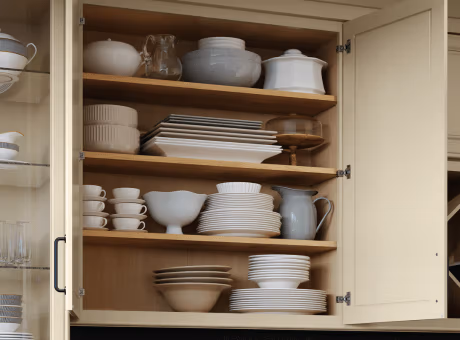 An open kitchen cabinet showing organized shelves filled with white and neutral-toned dishware, bowls, plates, and a jug.