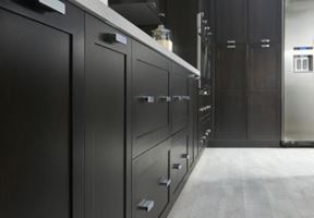 A view of a kitchen interior showing dark cabinets with sleek handles, light flooring, and part of stainless steel appliances.