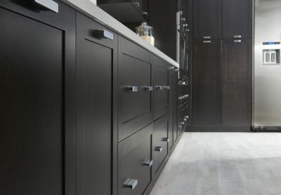 A view of a kitchen interior showing dark cabinets with sleek handles, light flooring, and part of stainless steel appliances.