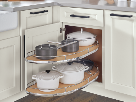 A white corner base cabinet with two pull-out trays storing cookware including Dutch ovens and saucepans. The trays are kidney-shaped with chrome rails and wood-look bottoms, optimizing storage in a corner.