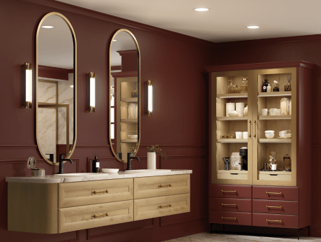 A luxurious bathroom with floating natural wood vanities, brass fixtures, double mirrors, and a dramatic deep red wall color. A coordinating linen tower includes glass-front cabinets and drawers.