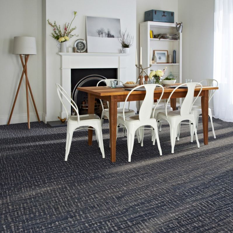 Dining room with a wooden table, white metal chairs, fireplace, and dark textured carpet flooring near a sunlit window.