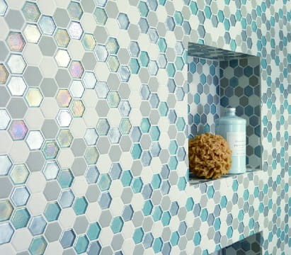 Close-up of a bathroom wall featuring blue, white, and aqua hexagon mosaic tiles with built-in shelving niches holding a sponge and bath product.