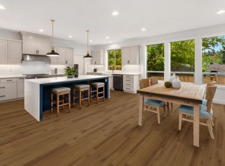 A modern kitchen interior with an island, bar stools, and a dining table in a bright, open space with windows overlooking greenery.
