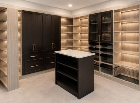A modern walk-in closet with a central island, shelves, and illuminated cabinets in a blend of dark and light tones.