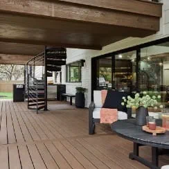 Covered outdoor patio with dark wood composite decking, black metal spiral staircase, black framed sliding glass doors, a black bench with an orange throw, and a round black table with drinks and flowers, overlooking a grassy backyard.