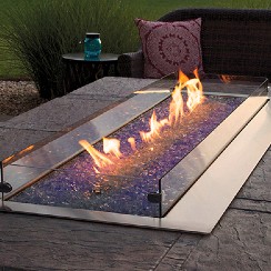 Rectangular outdoor fire table with blue fire glass and protective glass wind guards, set on a stone patio near outdoor seating.