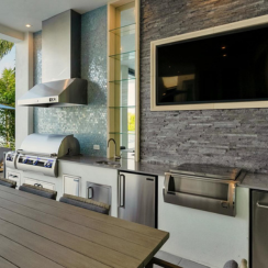 A luxury outdoor kitchen with a stainless steel built-in grill, illuminated control knobs, and matching accessories including storage drawers, a mini fridge, and a pull-out trash bin. A stainless steel side cooker, blue hexagon tile backsplash, and stacked stone wall with a mounted TV complete the sleek waterfront setup.