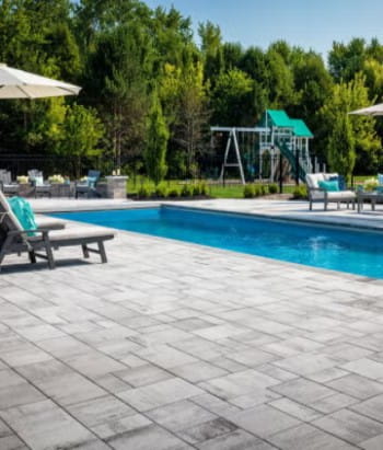 Outdoor pool area with gray stone paver patio, surrounded by lounge chairs, umbrellas, and a grassy yard with trees and a playset in the background.