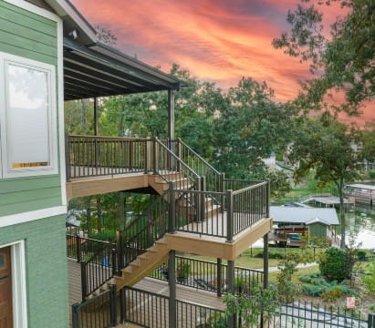 Two-story deck with black aluminum Key-Link railing and stairs overlooking a lake and trees at sunset, attached to a green house with white trim.