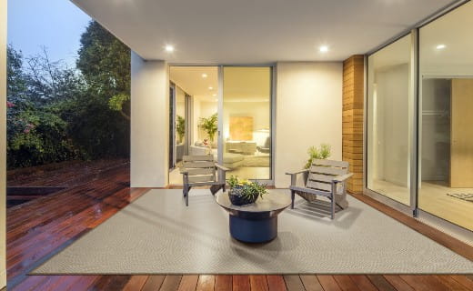 Modern covered patio with warm wood decking, a light gray outdoor area rug, two wooden chairs, and a round planter table, viewed from outside looking into a softly lit living room.