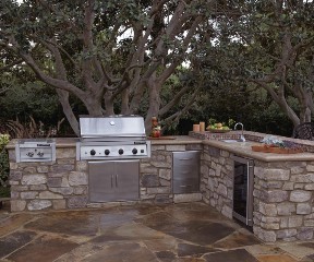 Outdoor kitchen with stone cabinetry, stainless steel grill, sink, mini fridge, and food prep area, set on a stone patio beneath mature trees.