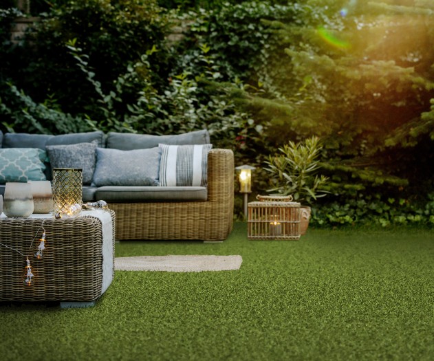 Cozy outdoor seating area with a wicker sofa and table, layered with pillows and lanterns, set on lush green artificial turf surrounded by greenery and soft sunset light.