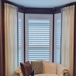 Armchair in front of bay windows with white blinds and cream curtains.