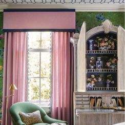 Pink drapes hanging over a window beside a green armchair and a rustic cabinet in a stylish room.