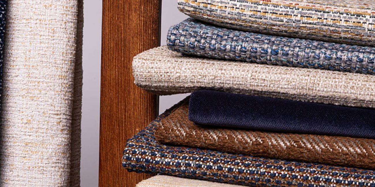Close-up of stacked upholstery fabric samples in neutral, blue, and brown tones next to a wooden frame.