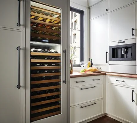 Modern kitchen with a wine fridge, wooden racks, a built-in coffee machine, and white cabinetry with black handles. A countertop with bread, wine, and olive oil under natural light.