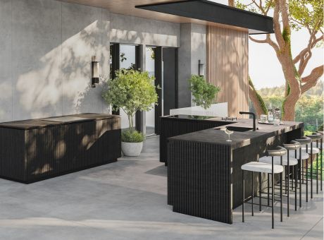 A modern open-air kitchen with counters, bar stools, and plants under a patio with light streaming through.