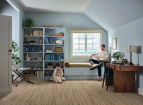 A study room with a desk, shelves, and a sitting area, where a person and child are present.
