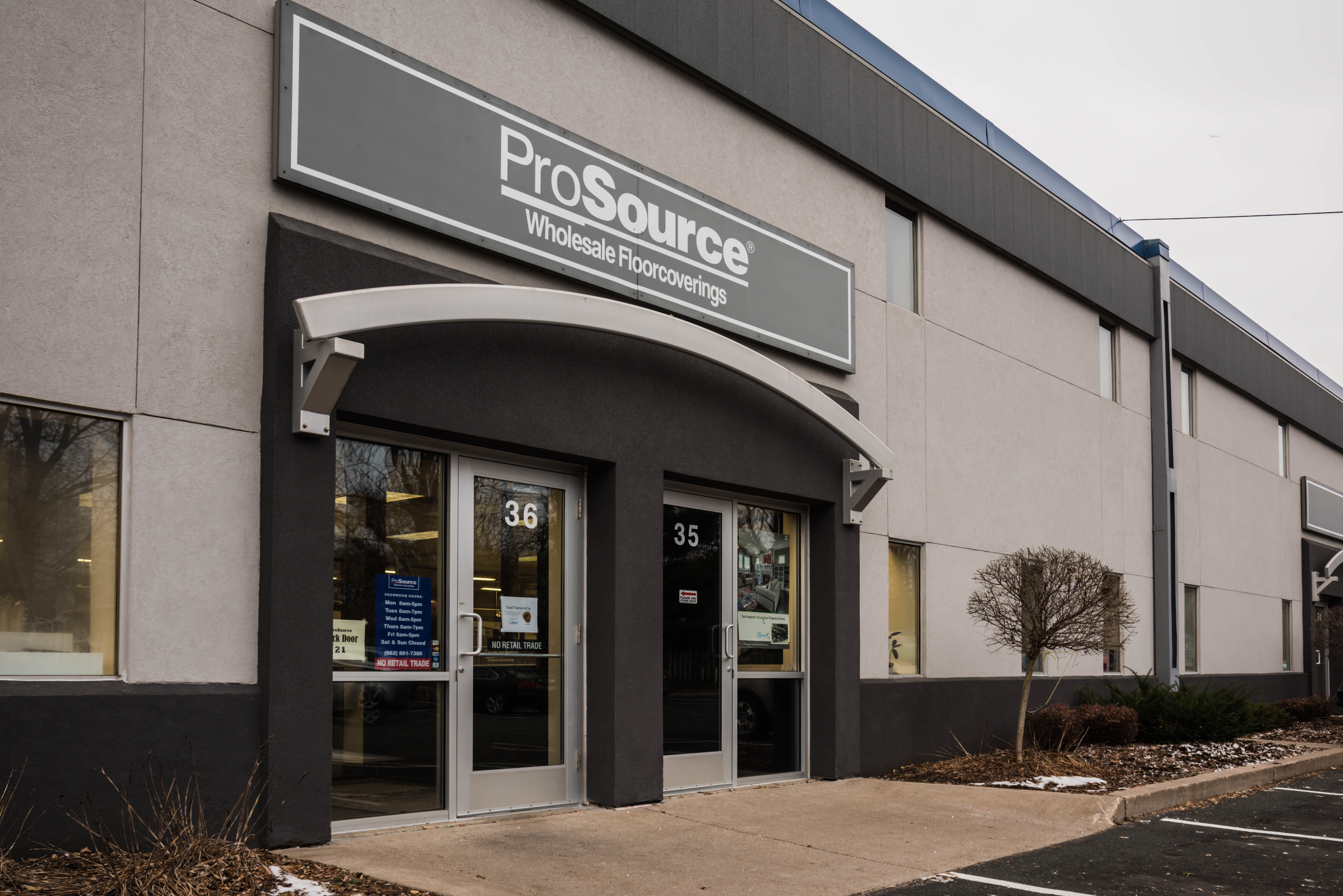 The exterior of a building labeled 'ProSource Wholesale Floorcoverings' with glass entrance doors and signage, featuring a parking area and landscaping.