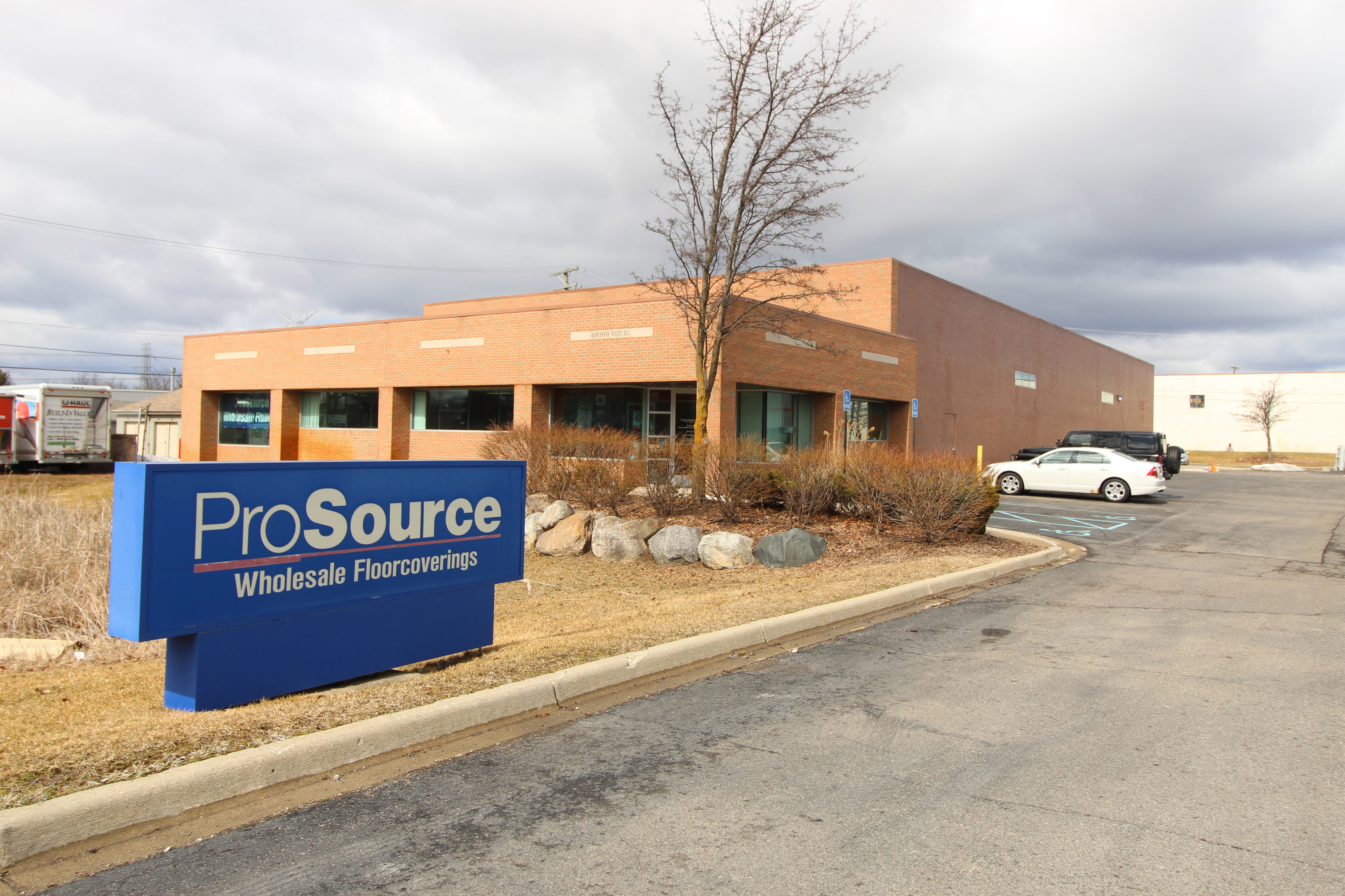 The exterior of the ProSource Wholesale Floorcoverings store featuring a blue company sign in the foreground and a brick building with glass windows in the background.