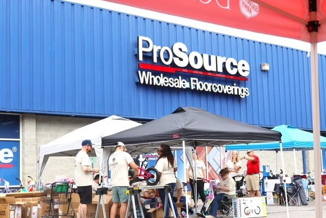 A community gathering outside the ProSource Wholesale Floorcoverings building, showing tents and people engaging in activities.