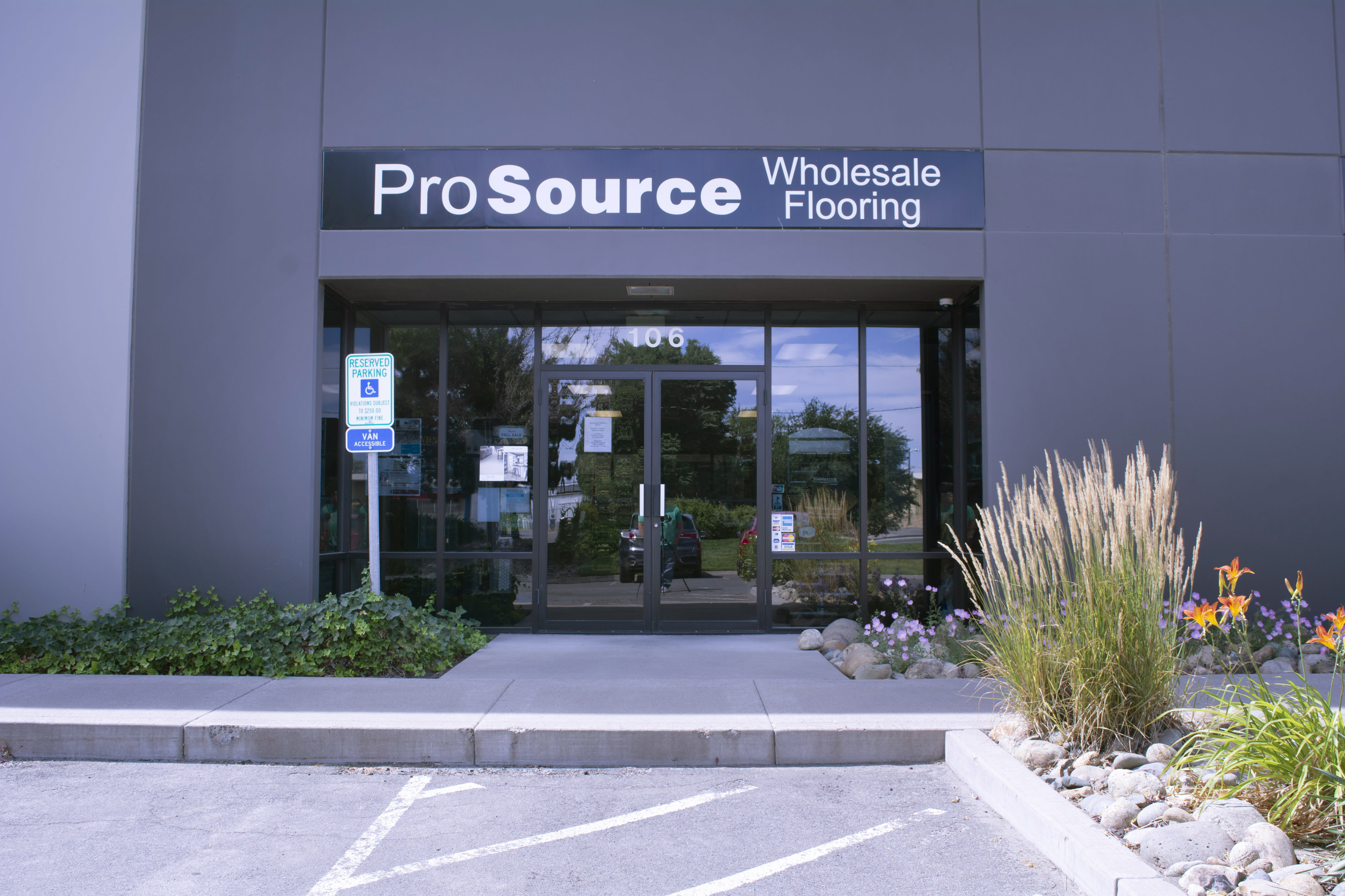 The entrance to a modern building marked as 'ProSource Wholesale Flooring' with landscaping and parking space visible.