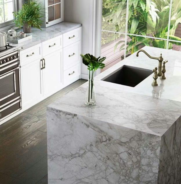 Modern kitchen with marble countertops, white cabinets, built-in sink, and outdoor greenery visible through a large window.