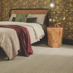 A bedroom interior with a bed, floral wallpaper, wooden side table, and beige carpet.