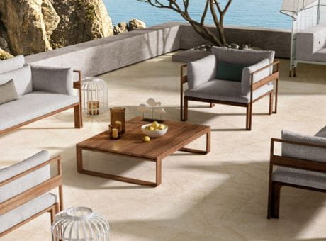 Outdoor patio setup with wooden and cushioned chairs, a wooden coffee table, placed on a light stone terrace near a waterfront and rocky background.