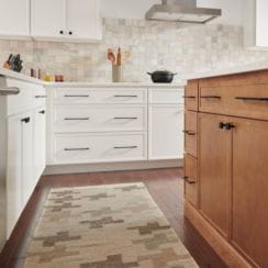 Interior view of a modern kitchen with white cabinets, wooden drawers, a patterned rug, and a tiled backsplash.