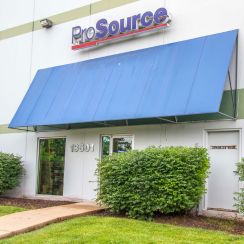 Storefront with ProSource sign and blue awning above the entrance, with greenery and a walkway in front.