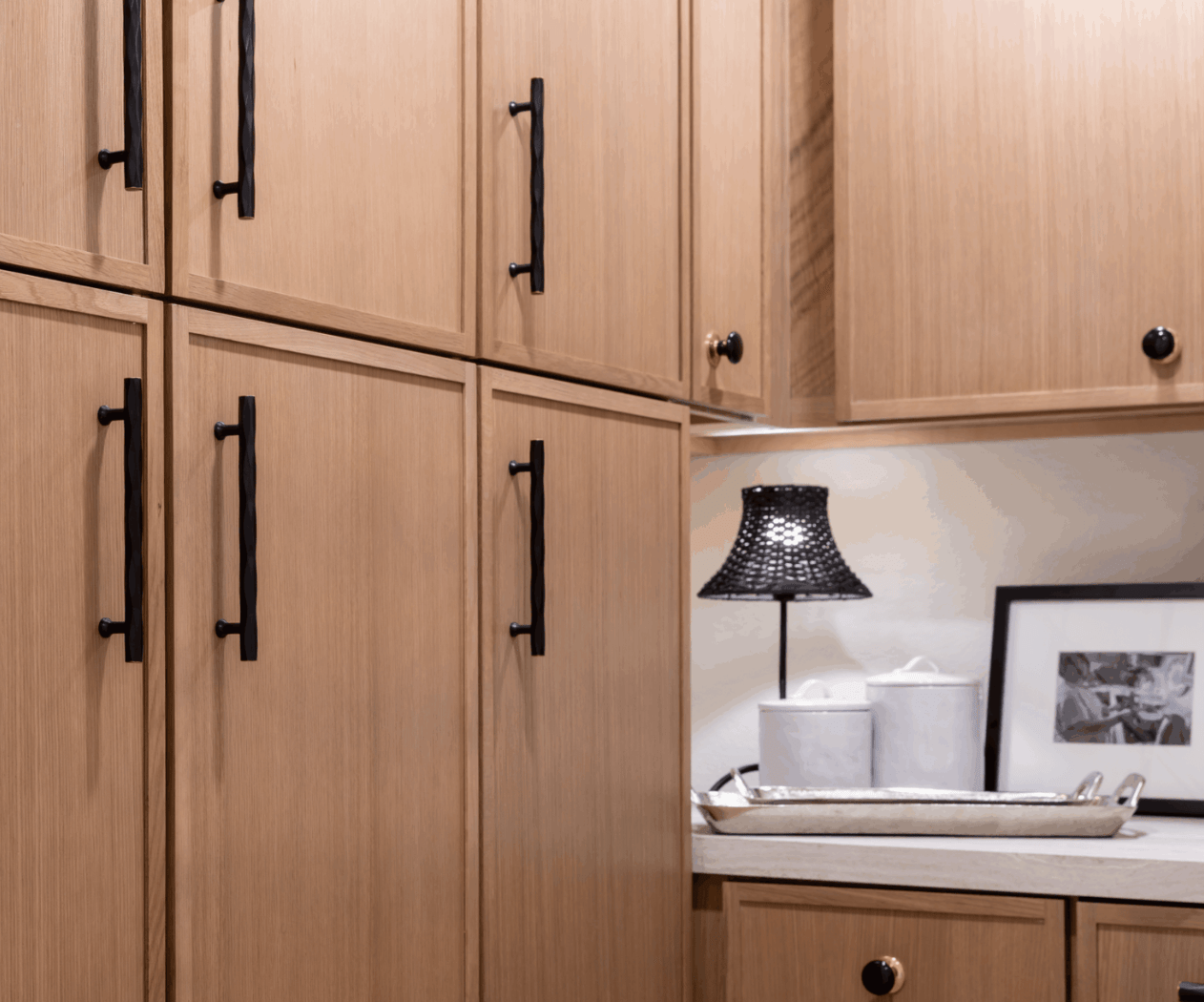 A close-up of wooden cabinets with black handles, a lamp with a black shade, and kitchen decor items on a countertop.