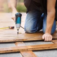 Person kneeling and using a power drill on a wooden surface.