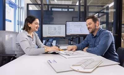 A man and woman sitting at a table, discussing architectural designs with visible monitors showcasing layouts and design materials on the table.