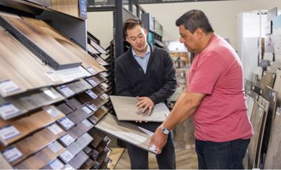 Two individuals in a retail store looking at flooring samples. One is showing a wooden plank sample while discussing details.